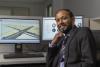 Angshuman Guin (a male professor wearing a black suit) sits at a desk in front of two monitors displaying data