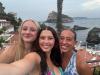 Three girls on a clifftop overlooking blue water taking a selfie.