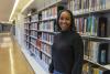 Person standing in a library aisle next to shelves filled with colorful books, wearing a dark sweater and jeans.
