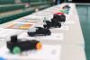 A row of small, student‑designed model race cars displayed on a table, each placed on top of design sheets in a gym setting.