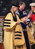 During a hooding ceremony indoors, a faculty member places a doctoral hood over the shoulders of a seated graduate wearing a gold gown and blue velvet doctoral regalia. Two additional faculty members in academic dress stand nearby, smiling. Rows of rolled diplomas are visible in the background.