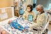 Two adults wearing protective gowns and gloves stand beside a hospital crib, using a tablet device while examining an infant lying on the mattress as medical equipment and monitors surround the crib.