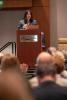 Vanessa Chan speaks at a podium at the Georgia Tech Hotel and Conference Center, addressing an audience. She holds a clicker and stands behind a laptop during a formal presentation.