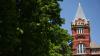 Georgia Tech’s historic Tech Tower rises above leafy green trees on a clear day, featuring a red brick facade, arched windows, and a pointed gray roof with the word “TECH” displayed prominently near the top.
