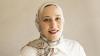 A professional studio portrait of a woman smiling warmly, wearing a light-colored patterned hijab and a white collared blouse against a neutral, solid background.