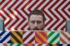 Artist Christopher Paul Dean peeks out from behind one of his artworks featuing crisscrossed lines of colors and chevron patterns.