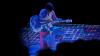 Against a black background, a woman in a silver jumpsuit sits on a chair playing a white guitar. Digital patterns in blue, purple and white are projected on her, the guitar, and the floor around her.