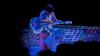 Against a black background, a woman in a silver jumpsuit sits on a chair playing a white guitar. Digital patterns in blue, purple and white are projected on her, the guitar, and the floor around her.