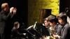 Four musicians are seated, playing saxophones and leaning towards the music stands in front of them. Facing them we can see the pianist in profile. Behind them is a brick wall illuminated in a fluorescent yellow.