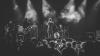 A black and white photograph of musicians on stage, smoke illuminated in the spotlights, the men playing guitar, bass, drums, and singing. Below and in front of them are concert-goers, looking up at the stage.