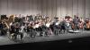 A full orchestra is arrayed on a stage, chairs in four rows stretching from edge to edge of the stage. 