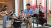 In a sunny room with floor to ceiling windows along one side, four musicians are playing electric guitar, drums, saxophone, and upright bass.