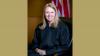 Formal portrait of Leigh May in judge's robes, set against a gold background.