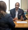 A man sits at a desk and is speaking to a woman. 