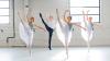 Four dancers are mid-movement, their right legs lifted and toes pointed towards the sky. The three women wear pale blue leotards with gauzy white skirts. The man wears a white T shirt and black tights.