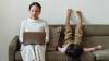 A sits on couch with a laptop with a young kid sitting next to them. 