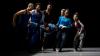 on a dark stage lit only from above and to the left, a group of seven dancers run in a semi circle. the middle five are carrying a prostrate body, another dancer.