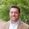 Portrait of Dr. Josh Eyler. Image is of a smiling man in a tan jacket in front of a natural green background. 