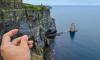 A hand holds a piece of rock with "GO JACKETS!" written on it in front of the Cliffs of Moher.
