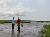Michael Hodges and Joel Kostka walk in the muddy marsh on wood planks with equipment used to determine marsh elevation. Marsh grass can be see in the background surrounding expansive mud flats.